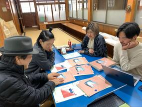 畳の部屋で資料を囲んで確認している様子。帽子をかぶった光島氏が指先で紙面を触りながら確認し、その様子を見守っている。