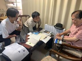 机を囲み、光島氏、今村氏、芝野氏の3人が資料を見ながら打ち合わせをしている様子。机の上にはさまざまな触図・触図つき会場地図が広げられている。光島氏が座ったまま１つの触図を触り、その様子を今村氏と芝野氏が確認している。